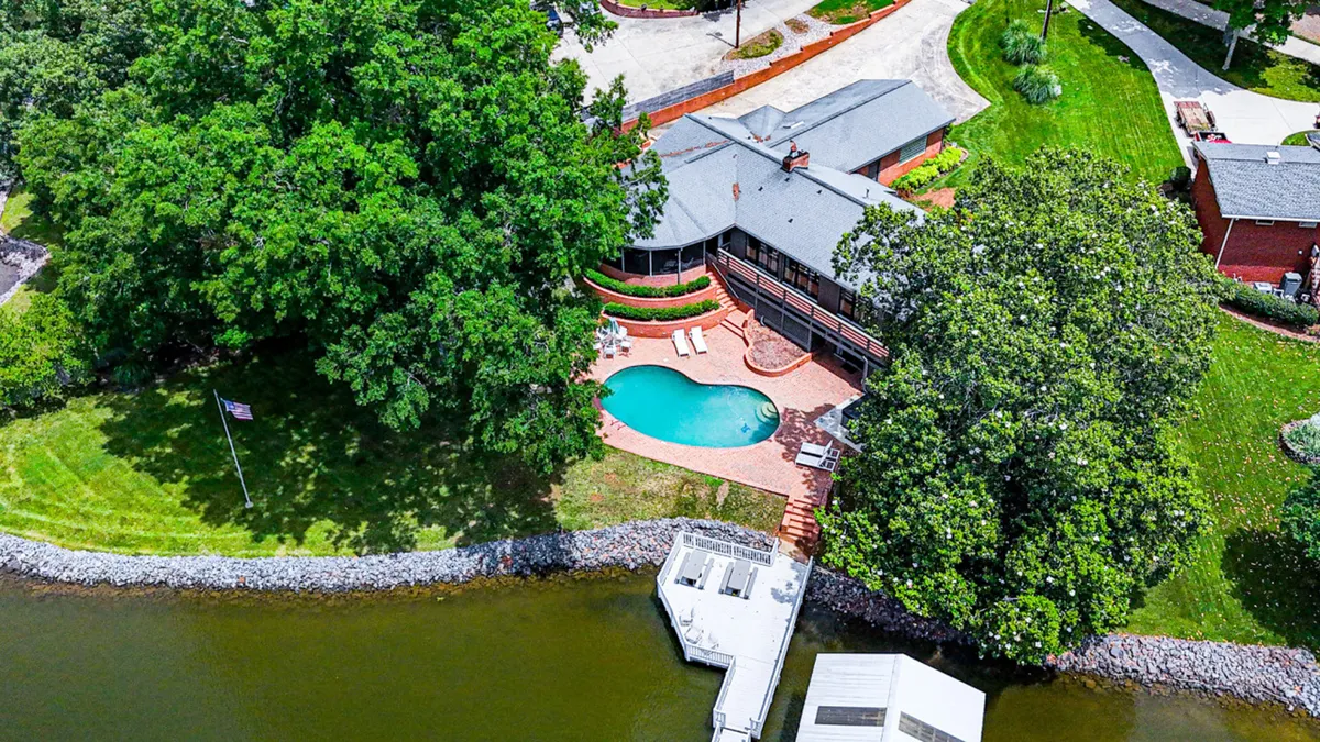 This exterior image showcases a sparkling swimming pool framed by large trees, with a deck area overlooking the expansive waterfront. The relaxed setting features ample space for swimming, sunbathing, fishing, and outdoor dining, and hints at sunset views over the water.