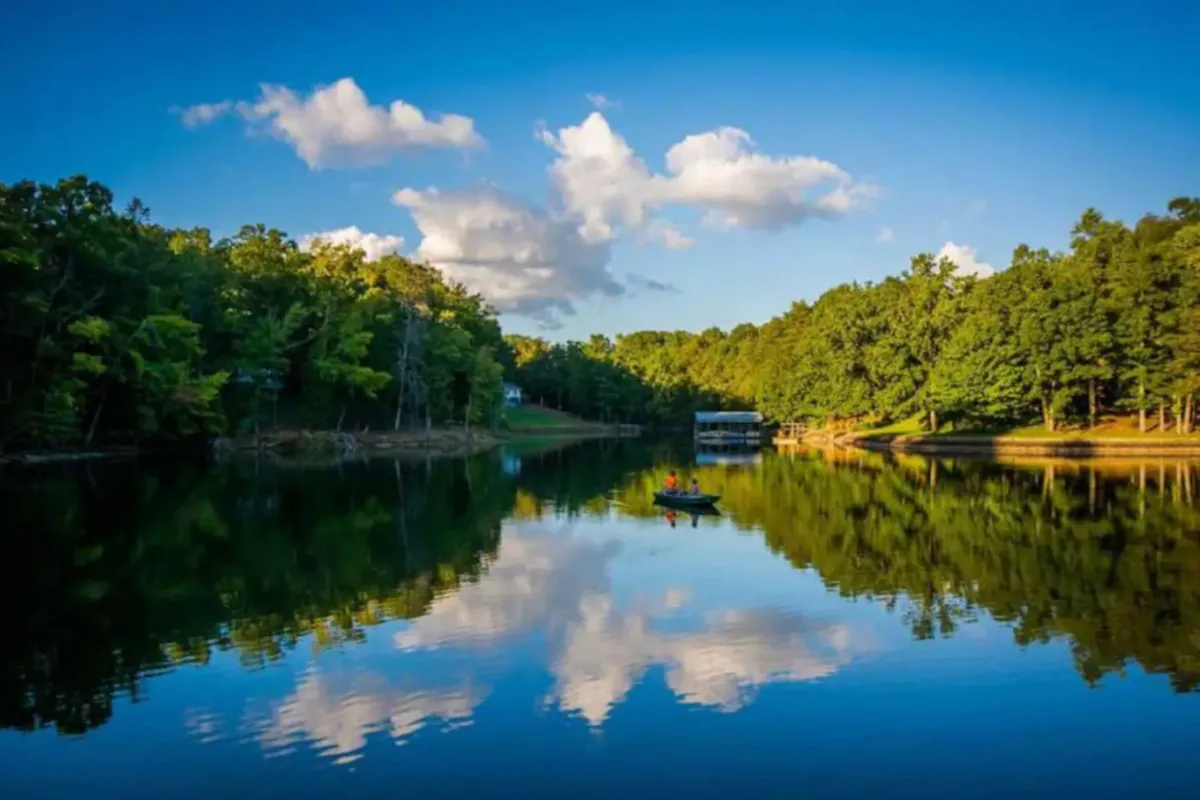 A broad, calm lake surface reflects lush green trees and soft clouds. Boaters glide across the water, and the untouched shoreline hints at both exploration and retreat. The natural beauty dominates the scene, emphasizing year-round outdoor possibilities.