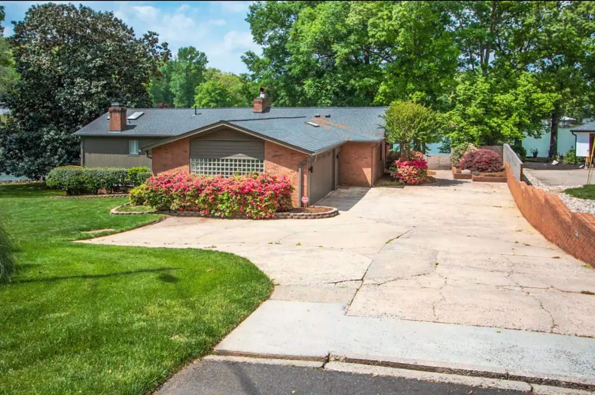 The brick exterior of the house is complemented by vibrant flower beds, a spacious driveway, and a lush, open green lawn. The setting is serene and welcoming, with clear spaces where children and pets can play and families can gather for outdoor activities.