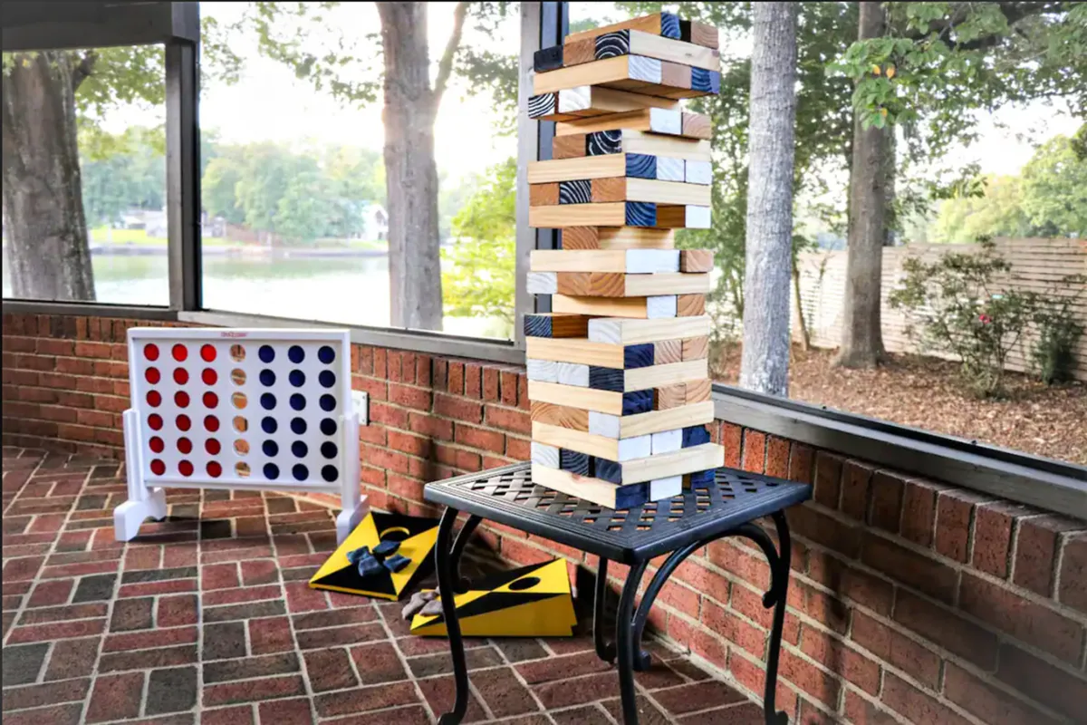 A cozy screened-in porch with plush seating is set up for a spirited game night, featuring a tall Jenga tower, Connect Four, and cornhole boards. Glass doors reveal outdoor views, blending the comfort of indoor living with the excitement of outdoor fun. Warm light and relaxed guests create an inviting, year-round atmosphere.