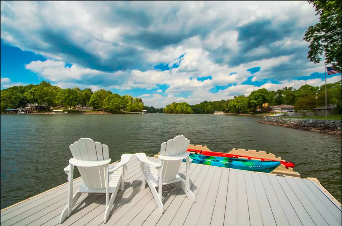 The image showcases a tranquil lakeside scene featuring classic Adirondack chairs set on a dock, with a vivid kayak nearby and calm Lake Wylie stretching out under a dramatic, cloudy sky. The atmosphere is serene, inviting guests to unwind by the water, launch a kayak for adventure, or simply soak in the peaceful lakeside setting.