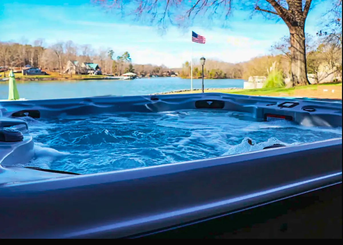 A tranquil hot tub set on a deck overlooking the lake, with an American flag and a tree nearby. The scene radiates peace and relaxation, making it an inviting spot for year-round soaking while enjoying panoramic lake views, whether at sunset or under the stars.