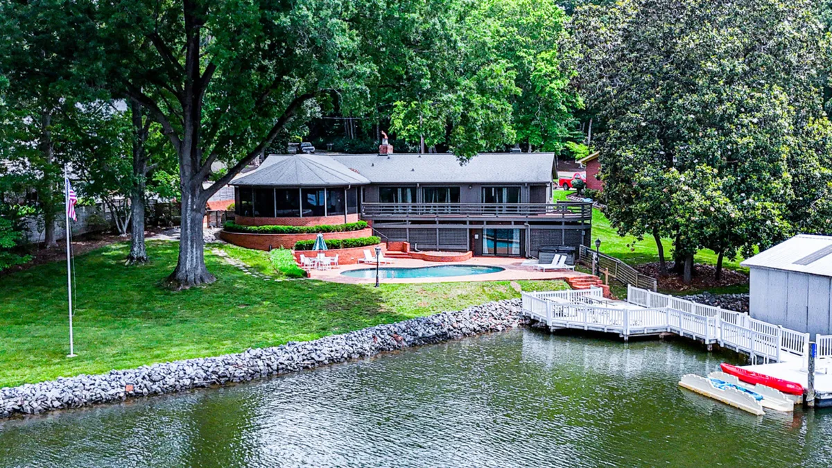 A wide exterior shot showcasing the house set beside the tranquil lake, with a large swimming pool, trees, a dock extending over the water, and a spacious deck area. The setting is ideal for swimming, boating, sunbathing, and family gatherings, capturing the property's all-in-one lakeside adventure vibe.