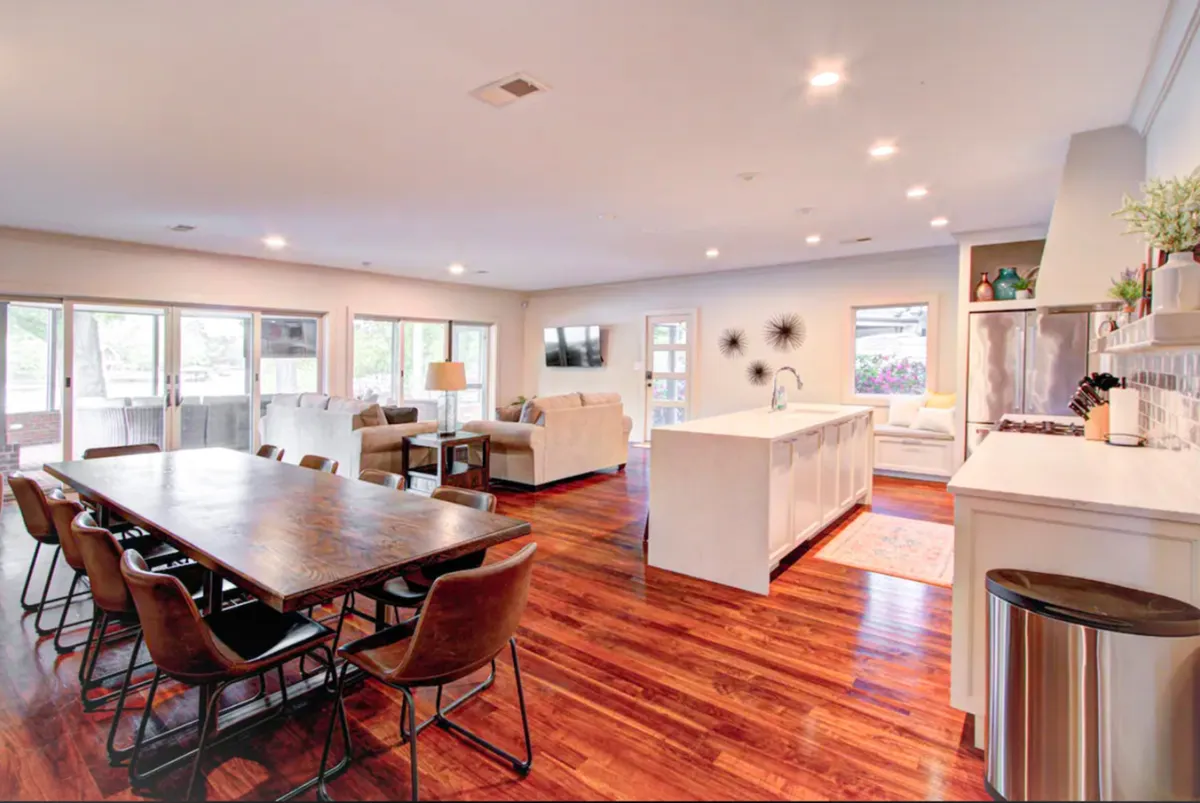 A sunlit open-concept space combining a large dining table, an island countertop, a comfortable seating area with plush sofas, and expansive windows offering stunning views of the lake. The area feels lively and welcoming, perfect for gatherings, shared meals, and relaxed conversations.