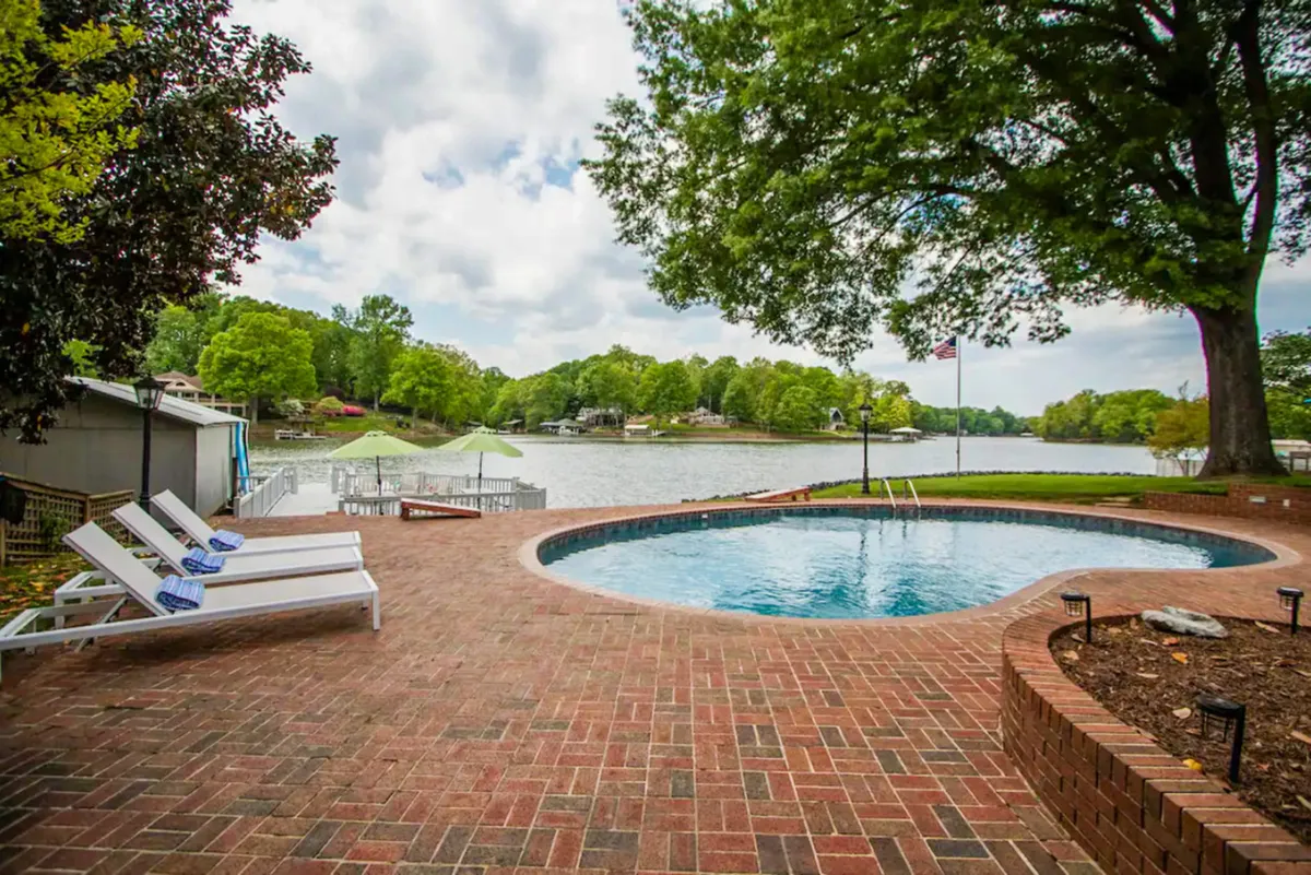 This image beautifully captures a serene lakeside pool setting framed by lush greenery and a spacious patio area. The tranquil water reflects the surrounding trees, and the scene exudes an atmosphere of leisure and exclusivity, perfect for both sunbathing and relaxed gatherings by the water.