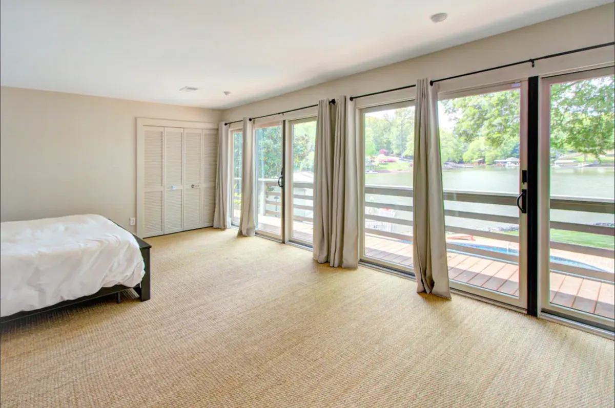 A serene bedroom featuring a minimalist bed placed near expansive windows that overlook the water. Natural textures in the rug and subtle, calming decor create a restful atmosphere, while the large windows flood the space with natural light and offer direct views of the lake.