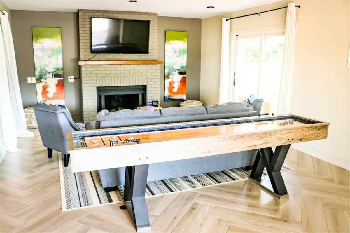 This interior image centers around a stylish shuffleboard table set within a welcoming room that includes a cozy sofa, fireplace, and wall art. The space feels perfect for relaxed games and socializing with friends or family. Large windows, if present, would support the sense of connection to the scenic outdoors.