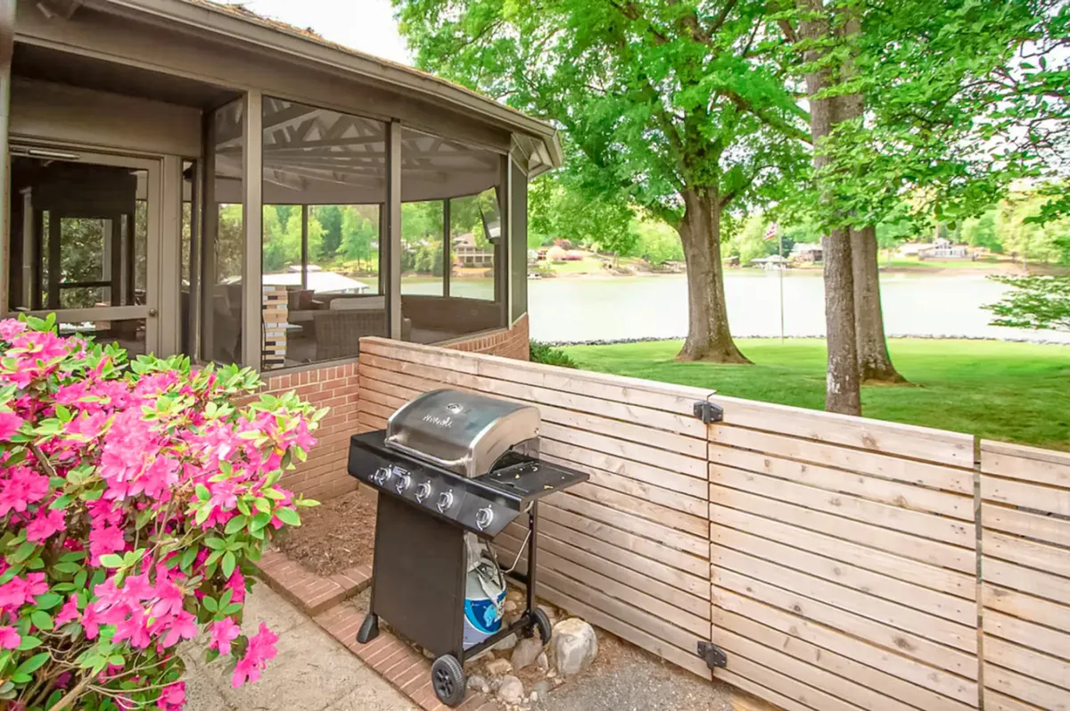 An inviting brick patio extends toward a screened-in porch, with vibrant flowering bushes and an expansive water view beyond. The grill and outdoor dining area suggest opportunities for alfresco meals, while the porch promises relaxation with uninterrupted sightlines to the lake.