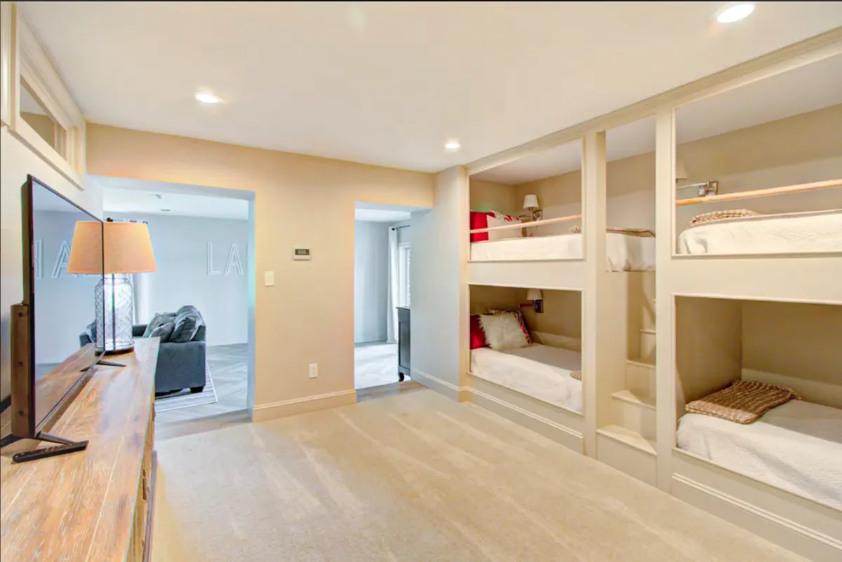 A cozy bunk room with four neatly arranged twin beds, a TV, and a lamp, designed for children or teens. The space feels playful yet comfortable, offering a dedicated retreat for younger guests to relax, play, or unwind together.