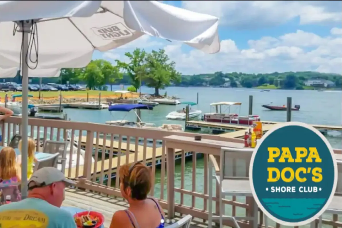 An outdoor dining area on a deck overlooking boats in the marina, with people visibly enjoying meals and socializing by the water. The mood is casual and cheerful, suggesting sunset dinners and vibrant group gatherings.