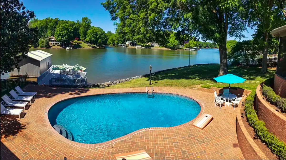 A wide shot of a sparkling swimming pool, outdoor seating area, and lush greenery, all overlooking a serene lake. The space combines areas for swimming, relaxation, dining, and enjoying nature, ideal for both pets and people to unwind after an active day.