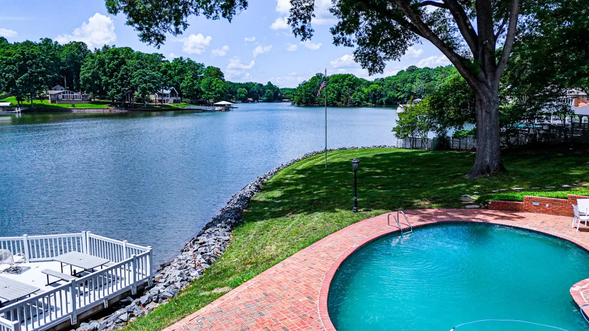 An inviting exterior scene highlighting lush green lawns, a shimmering swimming pool, mature trees, and a wide view of the lake. The space feels open and vibrant, ideal for pets to explore and for families to relax together outdoors. The overall atmosphere is relaxed, with plenty of space for play, picnics, and lakeside activities.